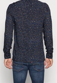 Mężczyzna ubrany w ciemnoniebieską koszulę z długim rękawem w czarno-brązowy wzór lamparta, połączoną z ciemnoniebieskimi dżinsami, widziany z tyłu.