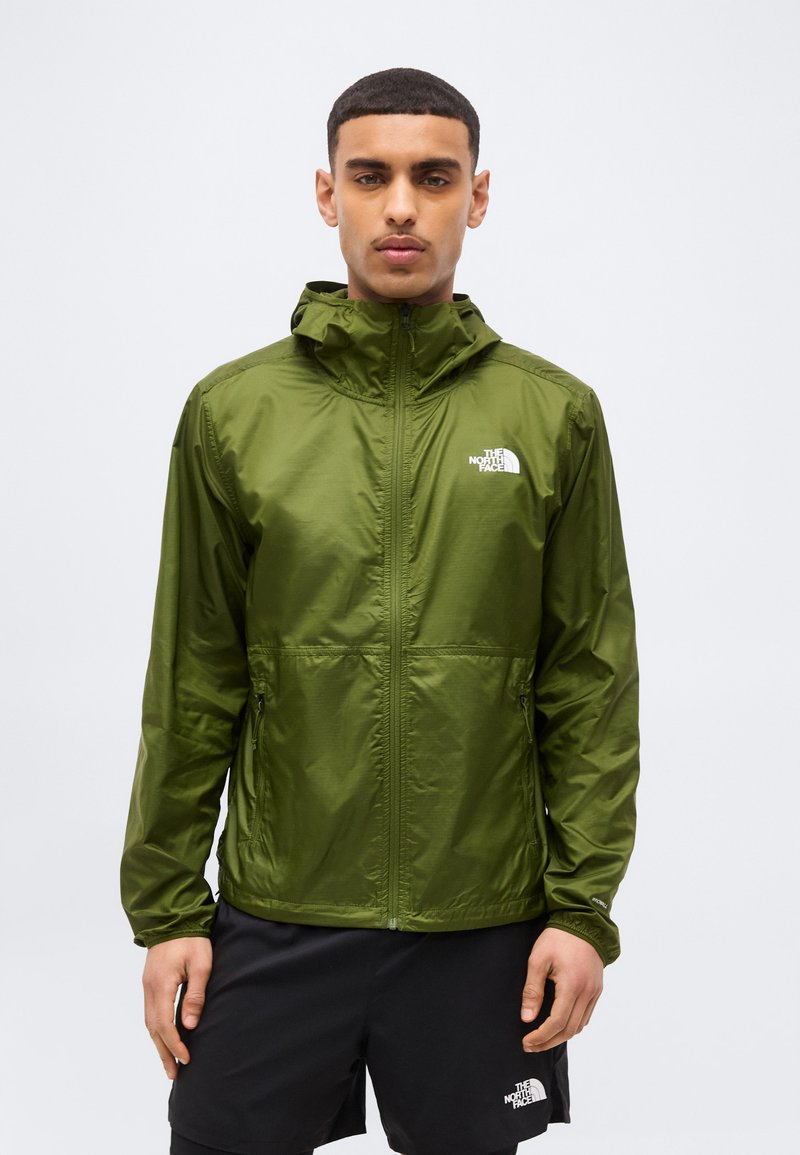 Ung mand iført olivengrøn North Face hættetrøje og sorte North Face shorts, stående mod en ensartet lys baggrund.