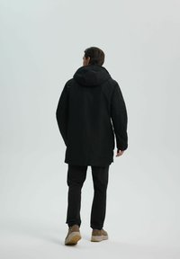 Parka nera impermeabile con cappuccio, caratterizzata da una linea dritta e maniche lunghe. Indossata con pantaloni neri abbinati e scarpe color marrone chiaro.