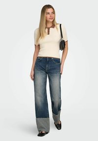 Femme portant un polo crème à manches courtes, un jean à jambes larges avec des patchs, des chaussures noires à bout carré et un sac à bandoulière noir, posant sur fond blanc.