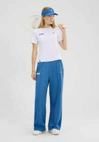 Camiseta atlética blanca con logo azul, combinada con pantalones anchos azules con bolsillos laterales. Raqueta de tenis sostenida en la mano derecha.
