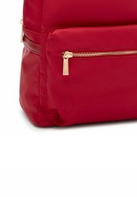 Angolo di uno zaino rosso in nylon con una tasca con cerniera dorata. Tessuto morbido, forma arrotondata e dettagli cuciti lungo i bordi.