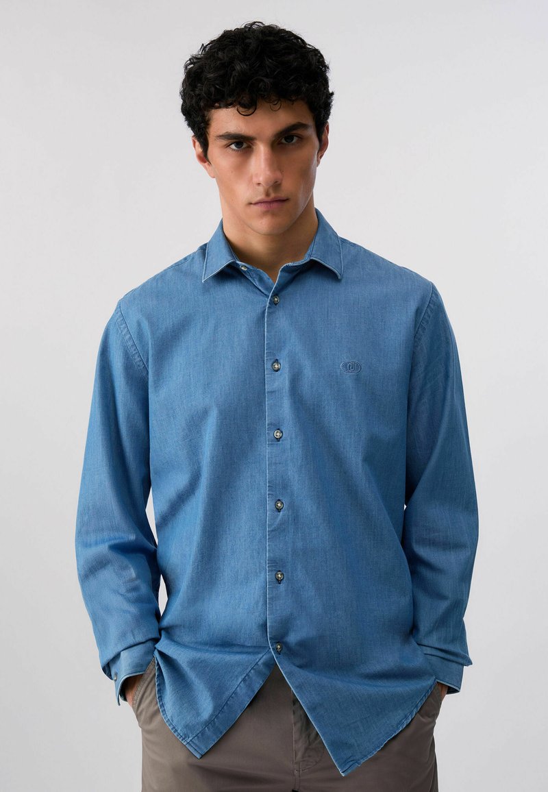 Jeune homme aux cheveux bouclés portant une chemise bleue à boutons et un pantalon beige, debout les mains dans les poches devant un fond uni.