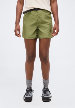 Persoană purtând pantaloni scurți verzi măsliniu cu curea, bluză bej, șosete gri până la gleznă și încălțăminte de drumeție neagră, stând pe un fundal alb.