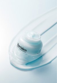 Torriden rauhoittava voide läpinäkyvässä purkissa, jossa on vaaleansininen kansi, ympäröitynä kiiltävällä voidejäljellä vaaleansinisellä taustalla.