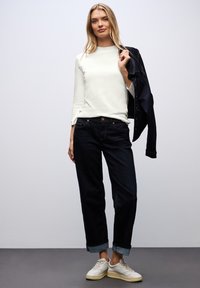 Camiseta blanca de manga larga, vaqueros de corte relajado en azul oscuro, zapatillas de color claro. Chaqueta colgada sobre el hombro; vaqueros remangados en el tobillo.