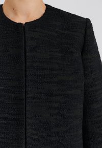 Černá texturovaná bunda s kulatým výstřihem a předním zapínáním, s kombinací tkaných vzorů a hladkých akcentů po celém povrchu.