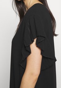 Femme portant un chemisier noir avec des manches volantes superposées et plissées, cheveux raides et foncés visibles sur un fond clair.