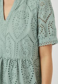 Svetlo zelena vezenina obleka z izrezom v obliki V, kratkimi napihnjenimi rokavi in očesnimi vzorci. Ima zapletene cvetlične vzorce in valovite robove.