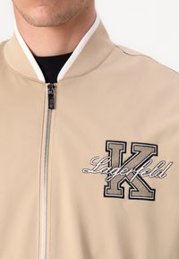 Beige bomber jacket with contrasting white ribbed collar, full zip, and embroidered "Lagerfeld K" logo in black and gold on the chest.