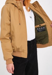 Personne portant une veste à capuche beige en train de dézipper pour montrer une doublure intérieure vert olive avec une étiquette d'entretien, un pantalon noir et une chemise blanche avec un petit logo.