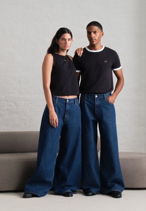 Une femme et un homme posent côte à côte, vêtus de hauts noirs, de jeans bleus à jambes larges et de chaussures noires, devant un mur de studio uni.