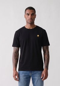 Άνδρας με τατουάζ που φοράει μαύρο t-shirt με μικρό σχέδιο χάμπουργκερ στο στήθος και μπλε τζιν, στέκεται μπροστά σε ένα απλό φόντο.