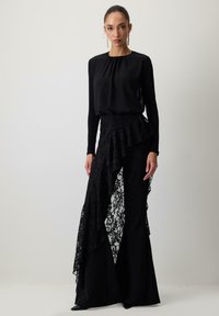 Melna virsma ar garām piedurknēm un savelktu kakla izgriezumu, apvienota ar slāņotu melnu mežģīņu svārku ar volānu apakšmalu un pielāgotām melnām biksēm.