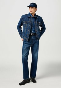 Giacca di jeans con due tasche sul petto, camicia blu scuro, jeans blu scuro abbinati e stivali neri alla caviglia. Indossato con un cappellino blu.