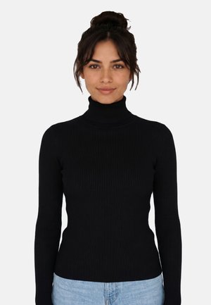 Donna con capelli scuri raccolti in uno chignon, che indossa un maglione nero a coste con collo alto e jeans azzurri chiari, in piedi davanti a uno sfondo chiaro e uniforme.