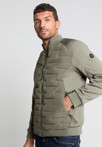 Groene gewatteerde jas met een hoge kraag, ritssluiting, zijzakken en textuurmouwen. Bevat een logo op de linkermouw.