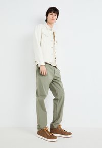 Chaqueta de mezclilla blanca, camiseta beige, pantalones verde claro, zapatillas altas marrones con cordones naranjas; fondo minimalista.