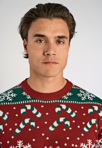 Punainen neulepusero, jossa on valkoisia lumihiutaleita ja vihreitä karkkitanko-kuvioita. Ribattu vihreä kaula-aukko ja teksturoitu pinta.