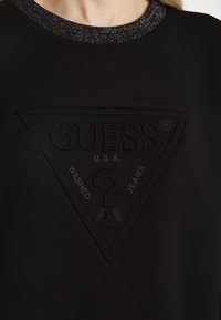 Sweat-shirt noir avec logo texturé Guess en forme de triangle, avec les inscriptions "U.S.A" et "Washed Jeans" sur la poitrine, porté par une personne.