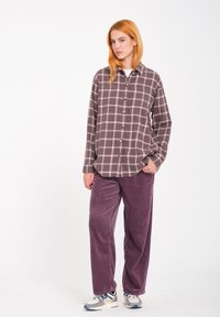 Camisa a cuadros en color malva con líneas crema, combinada con pantalones de pana en púrpura oscuro y zapatos estilo zapatillas. Textura de tela suave.