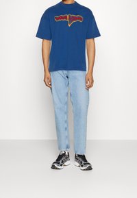Blaues Baumwoll-T-Shirt mit kurzen Ärmeln und einem bunten "Viva Light"-Grafikdesign, kombiniert mit hellblauen Straight-Leg-Jeans und schwarzen Sportschuhen.