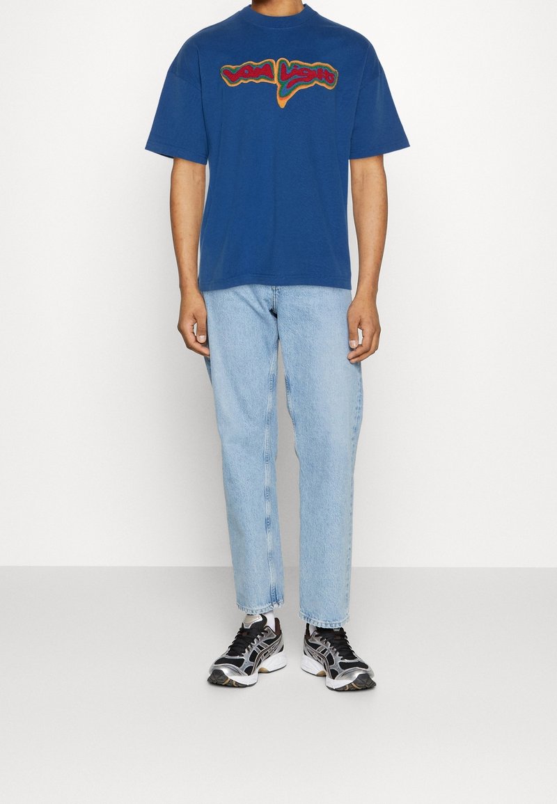 Blaues Baumwoll-T-Shirt mit kurzen Ärmeln und einem bunten "Viva Light"-Grafikdesign, kombiniert mit hellblauen Straight-Leg-Jeans und schwarzen Sportschuhen.