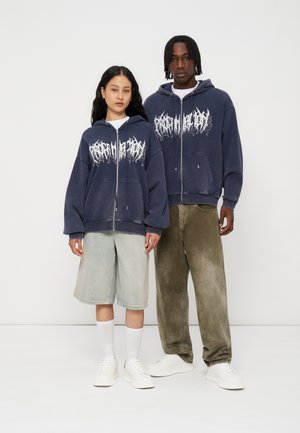 Two people wearing matching dark zip-up hoodies with white graphic text, paired with light shorts and dark pants, standing against a plain background.