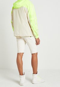 LEKKIE KURTKI Z KAPTUREM, Z NEONOWO ZIELONYMI RĘKAWAMI I KREMOWYM TYŁEM, W SPODNIKACH Z FRĘDZLAMI KOLORU BIAŁEGO I BUTAMI WYSOKIEJ CHOLEWKI.