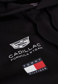 Svart stoff med hvit Cadillac Formula 1 Team-logo og rødt, hvitt og svart Tommy Hilfiger flaggmerke på klær.