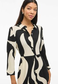 Črna in krema vzorčasta obleka s dolgimi rokavi, z gumbom spredaj in klasičnim ovratnikom; gladka tkanina in abstraktni oblikovni detajli.