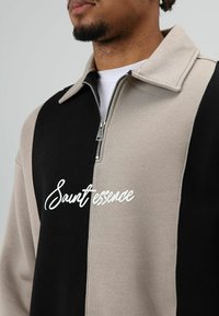 Quarter-zip pullover met zwarte en beige panelen, zachte stof en een wit cursief logo "Saintessence" op de borst.