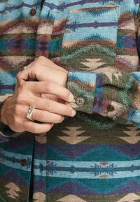 Overhemd met knoopsluiting, versierd met veelkleurige geometrische patronen in blauw, bruin en groen. Zachte textuur met zichtbare stiksels en twee voorzakken.