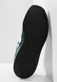 Dark green sneaker with a textured upper, white accents, and a rubber, hexagonal-patterned sole. The shoe is tilted for visibility.