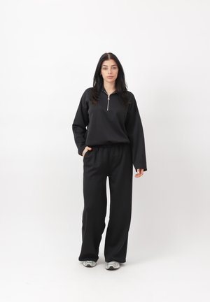 Femme aux cheveux foncés portant un haut noir à manches longues avec fermeture éclair, un pantalon noir ample et des baskets blanches, debout devant un fond blanc.