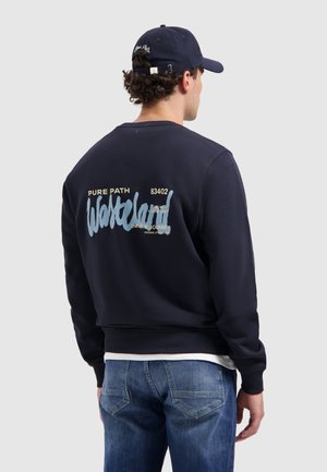 Mann trägt einen navyblauen Sweatshirt mit hellblauem und cremefarbenem Text auf dem Rücken, kombiniert mit blauen Jeans und einer navyblauen Mütze.