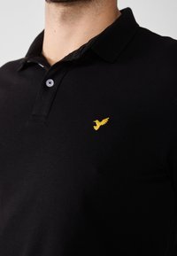 Črn polo majica, izdelana iz teksturirane tkanine. Ima rumeni izvezeni ptičji logotip na levem prsnem delu in ovratnik z dvema gumboma.