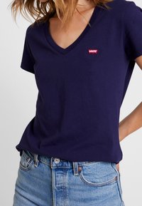 Mörkblå bomullströja med V-hals och korta ärmar. Har en röd Levi's logotyp på bröstet. Bärs med ljusblå jeans.