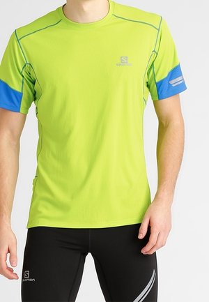 Mann trägt ein kurzärmeliges Sportshirt in Limettengrün mit blauen Ärmelakzenten und schwarze Sportleggings mit reflektierenden Details.