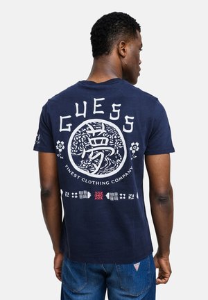 Hombre de espaldas vistiendo una camiseta Guess azul marino con logo y texto de inspiración asiática en blanco, combinada con vaqueros azul denim.