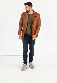 Bruine leren jas met een iets ruwe textuur, gesloten rits en borstzakken, gecombineerd met een groene shirt, donkere jeans en lichte sneakers.