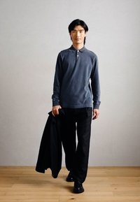 Πολυκαιρισμένο, ναυτικό μπλουζάκι με μακριά μανίκια και δύο κουμπιά, σε συνδυασμό με μαύρο παντελόνι. Άτομο κρατάει μια μαύρη ζακέτα. Ξύλινο δάπεδο και ουδέτερο φόντο.