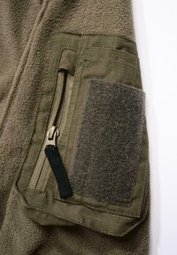 Olivengrønt fleece-stoff med en teksturert rektangulær lapp og detaljert glidelåslomme. Har svart nylon glidelåstrekk og rutenettdetaljer.