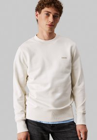 Sudadera crema con puños y dobladillo acanalados, con un pequeño logo marrón de Calvin Klein en el lado izquierdo del pecho, combinada con jeans oscuros.