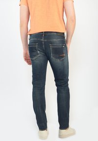 Jeans de mezclilla azul oscuro con un ajuste slim, que cuentan con bolsillos traseros, un ligero desgaste y un pequeño desgarrón cerca de los bordes de los bolsillos.