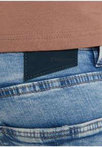 Närbild av blå denimjeans med en svart varumärkesetikett delvis synlig under en brun skjortslut.