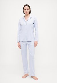 Ljusblått pyjamasset med knäppskjorta och långa byxor. Slät tyg, långa ärmar, sidfickor och pipingdetaljer vid manchetterna och kragen.