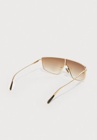 Γυαλιά ηλίου με χρυσή κορνίζα, φακούς με διαβάθμιση καφέ και λεπτούς βραχίονες που ακουμπούν σε λευκή επιφάνεια.