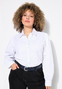 Camisa blanca de botones con un sutil patrón bordado, mangas largas abullonadas y cuello, combinada con pantalones oscuros y un cinturón azul.