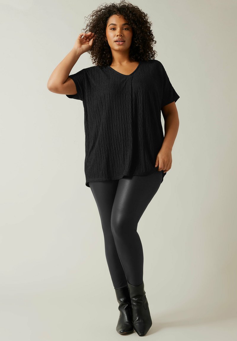 Schwarzes strukturiertes V-Ausschnitt-Oberteil mit kurzen Ärmeln, kombiniert mit schwarzen Leggings und Stiefeletten. Das Oberteil weist ein geometrisches Muster auf.